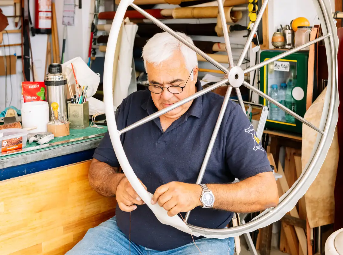 Artesano cosiendo a mano una funda de cuero sobre la rueda de timón en su taller, Port Ginesta